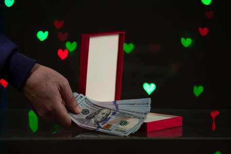 Valentine's Day gift. A hand puts a bundle of dollars in a red box. Behind on a dark background red...