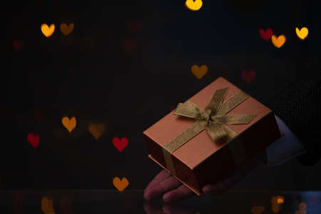 Valentine's Day gift. A hand in a jacket and white shirt holds a box with a bow. Behind on a dark background, multi-colored hearts glowの写真素材