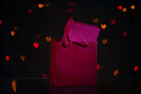 Valentine's Day. A gift in a red box is in a red paper bag. Behind on a dark background, multi-colored hearts glowの写真素材
