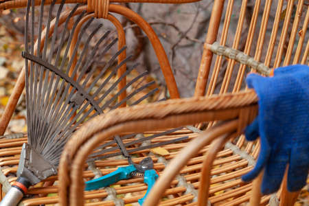 Autumn still life. A fan rake and garden shears lie on a beautiful wicker chair in an autumn park. Nearby are blue gloves. Close-upの写真素材