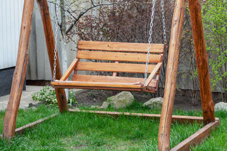 Cute brown wood swing hanging from chains on wooden poles mounted on a green grass lawn in the yardの写真素材
