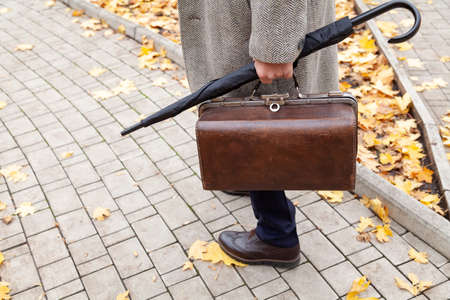 gentleman's walk. A man in stylish shoes and a white coat walks along the narrow alley of the autumn park. In the hand an umbrella-cane and a valiseの写真素材