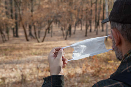 man in a cap holds a white medical mask in front of him in an autumn forest. Back view