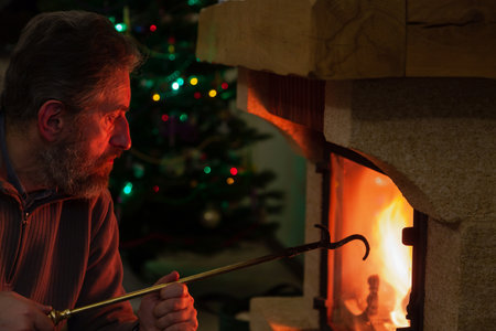 New Year Eve in front of the fireplace. The man looks at the fire in the burning fireplace. In the background, New Year lights are lit on the tree. Holds a brass poker in his handの写真素材