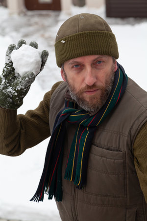 Snow games in the air. A man with a beard in a warm vest and a hat throws snowballs in the yard of a private house on a frosty dayの写真素材