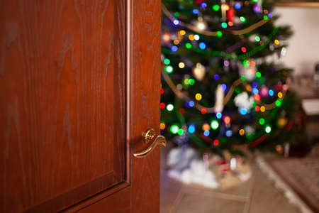 New Year comes into the house. Open brown wooden door with golden handicraft close-up. A decorated Christmas tree stands in the background in the room. Blurry garland lights are burning on the treeの写真素材