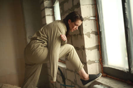 pretty woman in a gray jumpsuit stands in front of a large window at a construction site. A female portrait without retouching with her natural imperfections. The window is covered with polyethyleneの写真素材