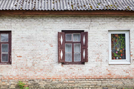 Ancient and modern nearby. The wall of a one-story house with a wooden window with open carved shutters. Next to it is a window with a white plastic frame and fresh flowersの写真素材