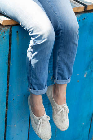 The slender legs of a woman hang from a wooden jetty made of blue wooden planks. White soft shoes on my feetの写真素材