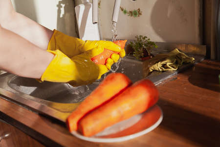 Hands in yellow rubber gloves wash a red carrot under running water. In the foreground are two defocused carrots.の写真素材