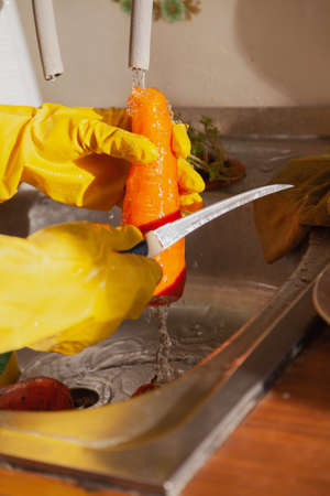Hands in yellow rubber gloves cut the carrot with a thin knife in the weight. Close-up. A stream of water flows on the rightの写真素材