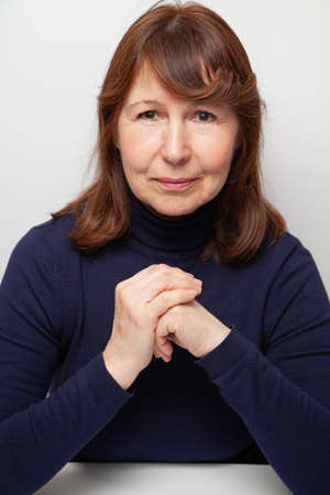 Portrait of a woman without retouching sitting on a light solid background. An elderly beautiful woman in a dark sweater looks into the frameの写真素材