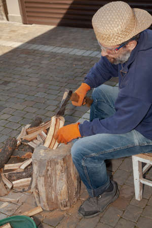 man with a beard sits in front of a tree stump and chops wood with an ax. He is wearing safety glasses and glovesの写真素材