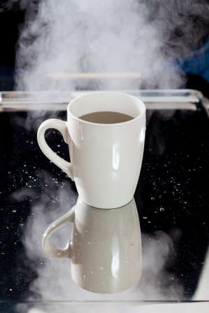 beige tall cup stands on a mirrored tray. Above the cup is a dense cloud of white vapor on a black...