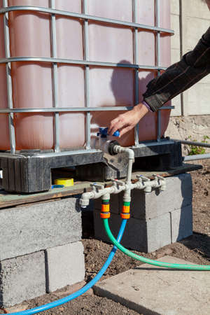 Irrigation system management. A hand opens a tap on the plastic pipes of the garden watering system. A container with water is installed above the pipesの写真素材