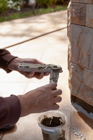 We install the tiles on the wall of the house. A hand applies a layer of special glue to flat natural stone tiles using a narrow trowel. Close-upの写真素材