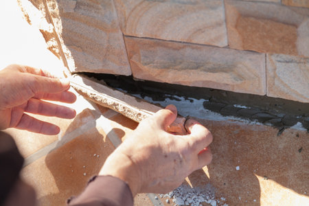 A worker hands insert a flat natural stone tile into the bottom row of the house plinth. Close-upの写真素材