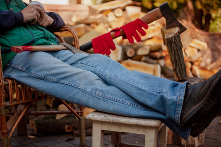 man is resting in a wicker chair in the yard in front of a pile of chopped wood. Outstretched legs in blue jeans lie on a wooden stoolの写真素材