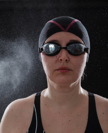 Portrait of a woman in a swimsuit on a black background. On his head are swimming goggles and a black cap. Small drops of water are visible in the backgroundの写真素材