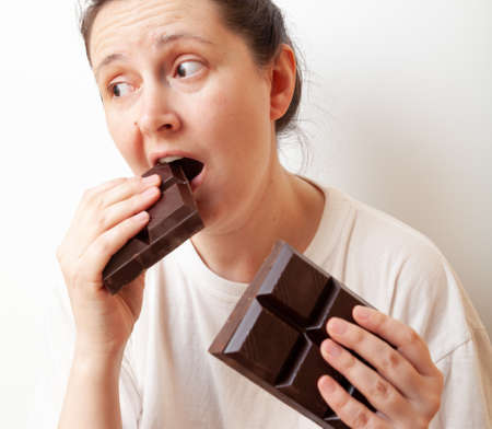 A sad looking young woman is biting half a large bar of dark chocolate. The second half of the tile...