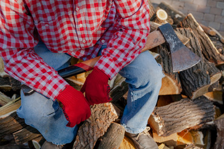man with a long-handled ax in red gloves sits on top of a pile of mulberry firewoodの写真素材
