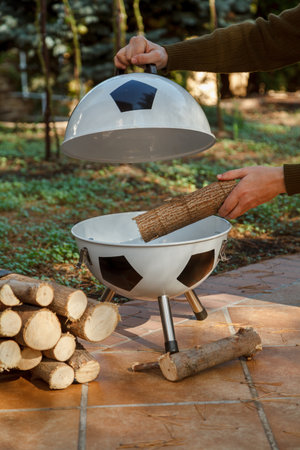 Football picnic. A hand puts a piece of wood into a new original brazier in the shape of a soccer ball. Nearby a bunch of firewoodの写真素材