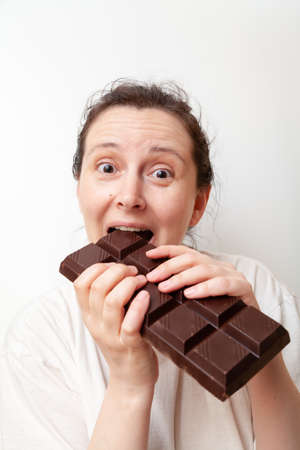 Young woman with a satisfied look bites a large bar of dark chocolate, holding it with two hands on a white backgroundの写真素材