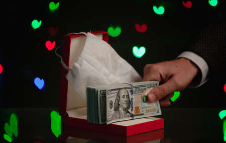 gift for Valentine's Day during the pandemic. A hand holds a bundle of dollars over a red box. Above is a medical mask. Behind on a dark background, multi-colored hearts glowの写真素材