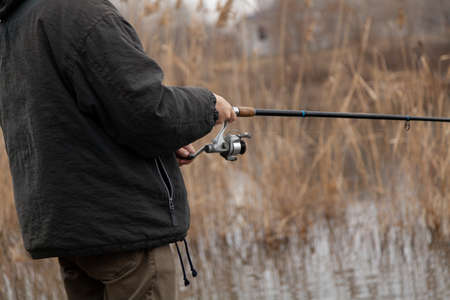 The hands of a spinner turn the handle of a spinning reel on a telescopic fishing rod on an autumn...