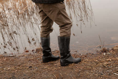 Legs in black rubber boots stand at the water's edge on the bank of the river close-upの写真素材