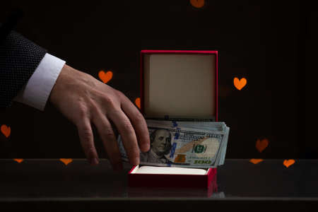 Valentine's Day gift. A hand puts a bundle of dollars in a red box. Yellow hearts are glowing behind on a dark backgroundの写真素材