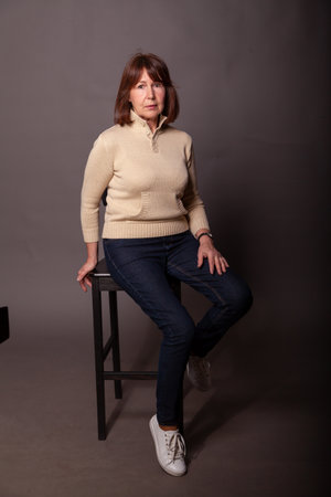 Portrait of a woman without retouching sitting on a dark plain background. Elderly beautiful woman in a beige sweater and jeans looks in the frameの写真素材