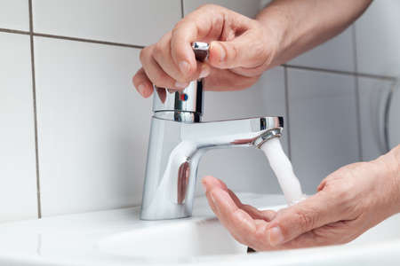 The right hand opens the tap on the lever faucet above the white tiled sink. A jet of water pours on the left hand. White tiles on the wallの写真素材