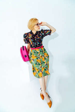 A stylish blonde woman with a short haircut in a skirt with a green floral print sits on a white background. Nearby is a pink bright bagの写真素材