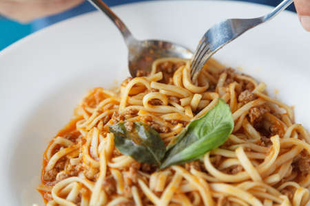 Hands put spaghetti with a spoon and fork on a round plate decorated with green leaves close-upの写真素材