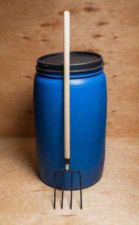 Fork and barrel. A blue round plastic container stands against a light-colored plywood backdrop. There is a black lid on top. A pitchfork on a wooden handle rests on a barrelの写真素材
