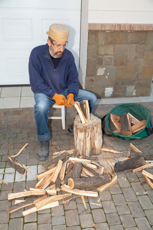 A man with a beard sits in front of a tree stump and chops wood with an ax. He is wearing safety glasses and glovesの写真素材