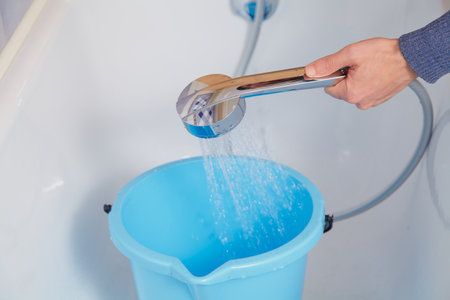 hand holds a shiny shower head and pours water into a blue plastic bucket. close-upの写真素材