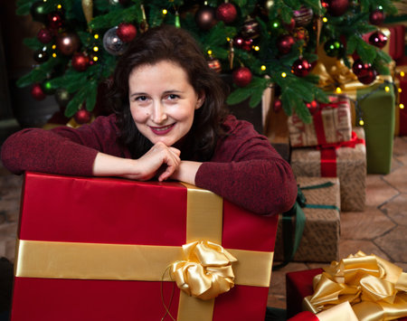 A beautiful young mother clutches a large New Year gift in a red festive package with a golden bow. In the background is a Christmas treeの写真素材