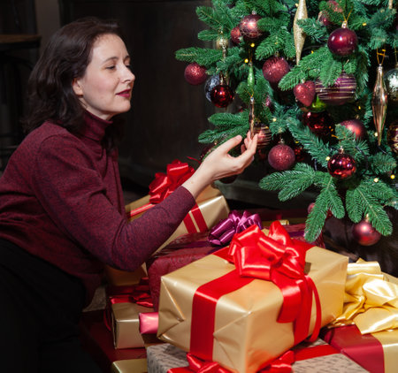 A contented young woman sits in front of a Christmas tree. There are a lot of boxes with gifts in festive packaging on the floor.の写真素材