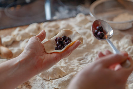 The hand puts a stuffing from black round berries in a dumpling spoon. On the table lie raw...