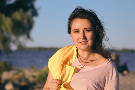 Portrait of a pretty girl with long red hair and a bright yellow jacket on the sandy beach of a wide riverの写真素材