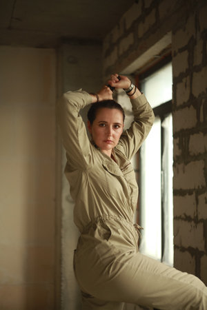 pretty woman in a gray jumpsuit stands in front of a large window at a construction site. A female portrait without retouching with her natural imperfections. The window is covered with polyethyleneの写真素材