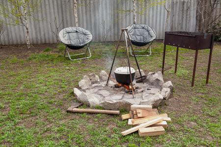 Equipped picnic area. A round stone campfire site is laid out in the meadow. The pot weighs on the counter. Nearby there is a brazier and two round armchairsの写真素材