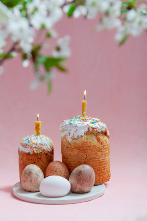 Easter still life. Two Easter cakes and eggs stand on a pink background. Burning candles are inserted into the cakes. Top view of a tree branch with white flowersの写真素材