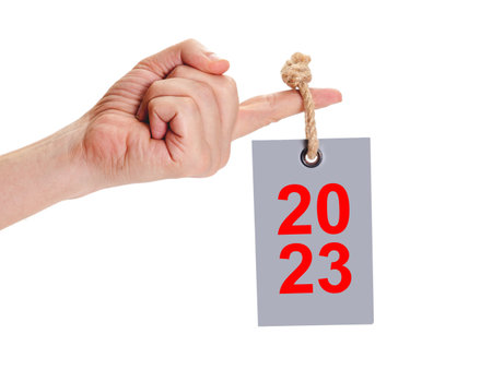 rectangular label hangs on index finger of left hand on white background. tag hangs on thick rope with large knot. tag reads 2023 in redの写真素材