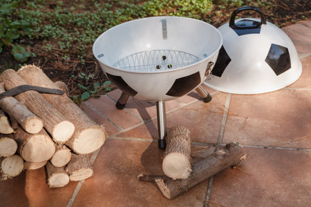 A soccer ball at a picnic. A new brazier in the shape of a soccer ball with a lid and a grate stands on a tile in the courtyard. Nearby a bunch of firewoodの写真素材