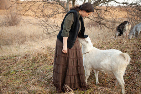 A shepherd woman in the countryside is herding goats in the autumn expansions of nature. The white goat wants tenderness and hides its head under the mistress's vestの写真素材