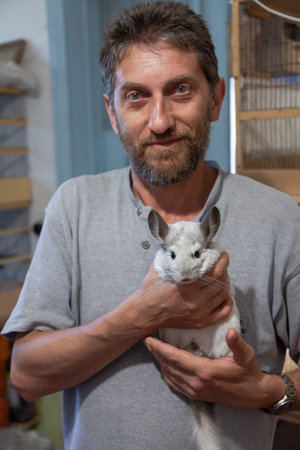 A zoologist with a beard hugs a contented white chinchilla. Cages with decorative birds are visible in the backgroundの写真素材