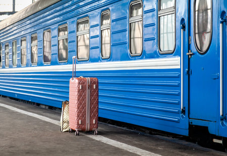 A modern suitcase on wheels with a long handle stands on an empty platform in front of a blue carriageの写真素材